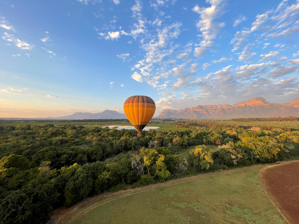 This image is of a Hot Air Balloon Flying over the Riverine Forest and Blyde River. There is lots of blue sky and white clouds visible.
